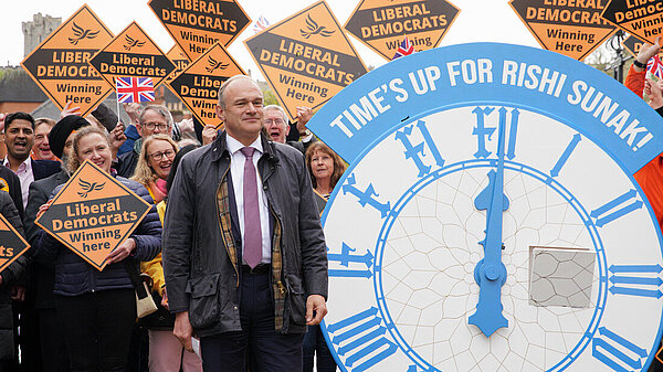 Ed Davey in front of crowd with Lib Dem diamond signs and clock with words time's up for Rishi Sunak